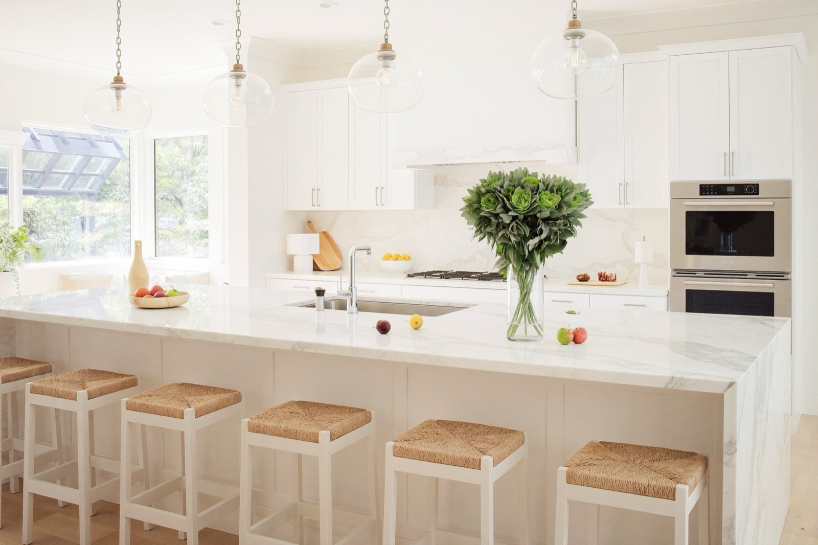 A white kitchen with a marble island, rush-seat stools and morning light through the windows.