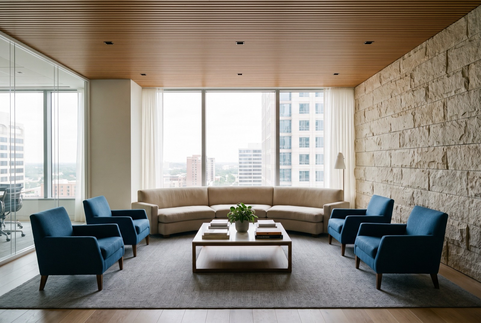 A high-floor lounge with blue armchairs, a stone wall and a city view through full-height glass.